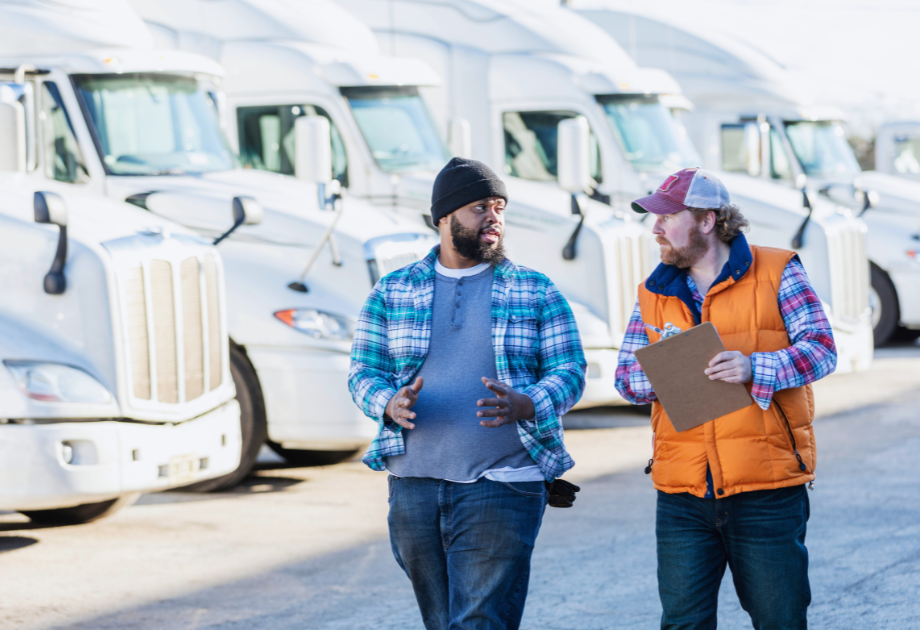 Two workers walking and talking in a truck yard with parked semi-trucks in the background.